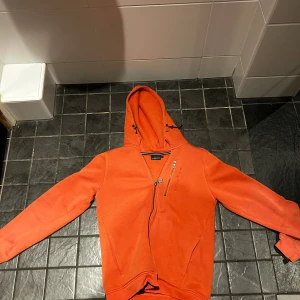Orange hoodie från Sail Racing - Säljer en orange hoodie från Sail Racing med huva och dragkedja framtill. Tröjan har en extra bröstficka med dragkedja och broderad logga på framsidan. Mjuk insida och ribbade muddar vid ärmslut och nederkant. Perfekt för dig som gillar sportig stil.