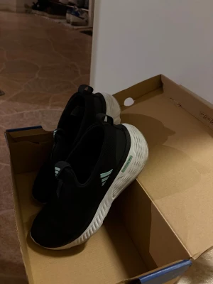 Svarta Adidas sneakers med vit sula - Snygga svarta Adidas sneakers med vit, vågformad sula och mintgröna detaljer på sidan. Skorna har en sportig design och är tillverkade i mesh och syntetmaterial för extra komfort. Perfekta för dig som gillar stilrena och moderna träningsskor.