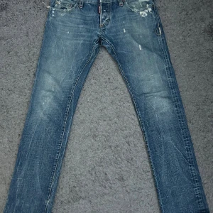 Dsquared2 jeans med slitningar - Säljer ett par blå skinny jeans från Dsquared2 med slitningar och distressed detaljer. Jeansen har klassisk femficksdesign, broderad röd lönnlöv på bakfickan och snygga slitna partier framtill och baktill. Perfekt för dig som gillar streetstyle och edgy look.