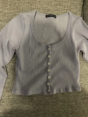 Ljuslila ribbad topp från Brandy Melville - Säljer en ljuslila ribbad topp från Brandy Melville med lång ärm och croppad passform. Toppen har rundad halsringning och knappar framtill. Perfekt för dig som gillar stilrena och trendiga plagg med enkel design.