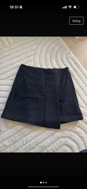 Svart omlottkjol från Stylein - Stilren svart kortkjol från Stylein i omlottmodell. Kjolen har en enkel och clean look med rak passform och är tillverkad i ett mjukt tyg som känns lite som ull. Perfekt att matcha med boots eller sneakers för en cool vibe.