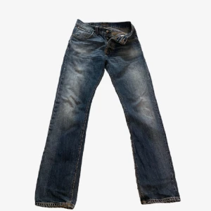 Nudie jeans Average Joe  - Snygga blå jeans från Nudie i modellen Average Joe med en skön passform och klassisk femficksdesign. Jeansen har slitningar och ljusare partier framtill för en cool, använd look. Tillverkade i 100% bomull och har orange kontrastsömmar samt broderade bakfickor.