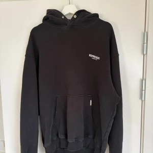 Svart hoodie Represent Owners' Club - Svart oversized hoodie från Represent med vit Owners' Club-tryck på ryggen och mindre logga på bröstet. Klassisk känguruficka, huva med tryckknappar och ribbade muddar. Perfekt för en streetwear-look.