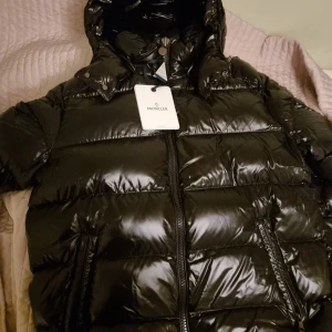 Svart Moncler Maya pufferjacka med huva  - Säljer en svart pufferjacka från Moncler Maya med glansig finish. Jackan har dragkedja framtill, två sidofickor med dragkedja och en ficka med Moncler-logga på ärmen. Perfekt för kalla dagar och har en riktigt snygg streetstil.