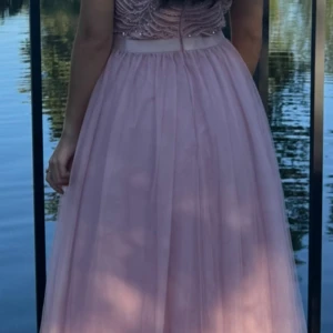 Ljusrosa långklänning med pärlor - En drömmig ljusrosa långklänning med transparent tyllkjol och pärlbroderad överdel. Klänningen har korta ärmar, rund halsringning och markerad midja. Perfekt för bal eller student. Materialet är lätt och ger en svävande känsla.