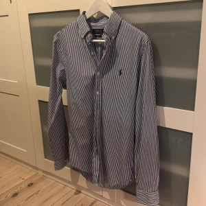 Randig skjorta från Polo Ralph Lauren - Snygg blåvit randig skjorta från Polo Ralph Lauren i slim fit-modell. Oxford krage, långa ärmar och knappar framtill. 