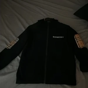 Svart Burberry vindjacka med huva - Svart vindjacka från Burberry med klassiska rutor i beige och brunt på ärmarna och insidan av huvan. Jackan har dragkedja framtill, huva och Burberry-logga på bröstet. Perfekt för dig som gillar stilrena detaljer och ikoniskt mönster.