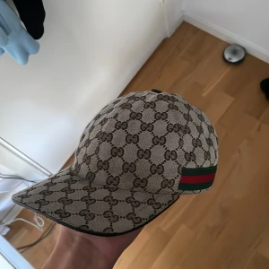 Gucci keps beige - Snygg beige och brun keps från Gucci med klassiskt GG-monogram över hela kepsen. På sidan finns de ikoniska gröna och röda ränderna. Justerbar rem baktill i svart. Materialet är slitstark bomull med detaljer i skinn. Perfekt för dig som vill ha en lyxig streetstyle.