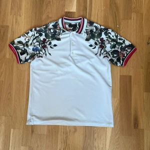 Zara skjorta  - Vit skjorta från Zara med blommigt mönster på axlar och ärmar. Kragen och ärmsluten har röda och vita ränder som ger en snygg kontrast. Skjortan har korta ärmar och knappar framtill. Perfekt för dig som gillar unika detaljer och stil.