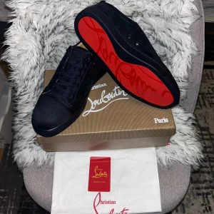 Svarta sneakers från Christian Louboutin - Säljer ett par svarta sneakers från Christian Louboutin med ikonisk röd sula och snygg snörning. Skorna har rund tå och är tillverkade i mocka med diskret broderad logga på hälen. Perfekt för dig som gillar exklusiv och stilren design.