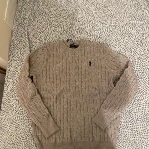 Beige stickad tröja Polo Ralph Lauren - Snygg beige stickad tröja från Polo Ralph Lauren med klassiskt kabelmönster och svart broderad logga på bröstet. Rund hals och ribbade muddar vid ärmslut och nederkant. Perfekt för dig som gillar stilrena och tidlösa plagg.