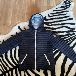 Mörkblå Colmar pufferjacka med huva - Snygg mörkblå pufferjacka från Colmar med huva och vita dragkedjor. Jackan har quiltad design, två fickor med dragkedja och Colmar-logga på ärmen. Insidan är ljusgrå och materialet är syntet. Perfekt för kalla dagar och riktigt stilren look.