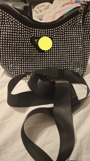 Svart axelväska med silverdetaljer - Cool svart axelväska med glittrande silverfärgade nitar på framsidan. Väskan har bred axelrem, dragkedja upptill och extra fack med dragkedja på baksidan. Materialet är skinnimitation och insidan är helsvart.