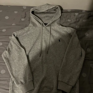 Grå hoodie från Polo Ralph Lauren - Snygg grå hoodie från Polo Ralph Lauren med klassisk broderad logga på bröstet. Tröjan har huva med snörning och ribbade muddar vid ärmslut och nederkant. Perfekt för chill dagar och enkel att matcha med jeans eller joggers.
