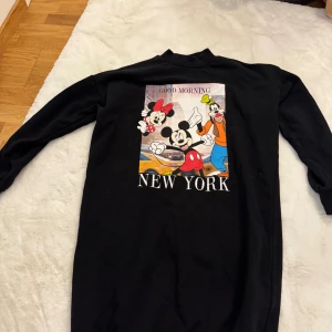Svart Disney klänning med tryck - Svart långärmad klänning från FB Sister x Disney i storlek M. Framsidan har ett stort färgglatt tryck med Mimmi, Musse och Långben samt texten 'Good Morning New York'. Mjuk bomullskvalitet och ribbad hals. Perfekt för dig som älskar Disney och streetstyle.