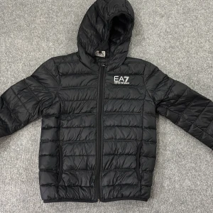 Svart pufferjacka från EA7 Emporio Armani - Svart pufferjacka från EA7 Emporio Armani i storlek 12A/ 150 Cm. Med huva och diskret logga på bröstet. Jackan har quiltad design, dragkedja framtill och två sidofickor. Perfekt för dig som gillar stilren och sportig look. Passar dig som vill ha en lätt och varm jacka med snygg passform. Jackan är använd 2 gånger. 