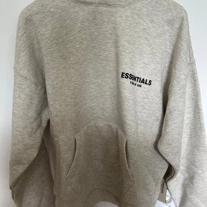 Helt Ny Grå Essentials Fear of God hoodie - Helt ny snygg ljusgrå hoodie från Fear of God Essentials med svart logga på bröstet och ryggen. Tröjan har huva, känguruficka och en relaxed fit. Perfekt för dig som gillar streetwear och vill ha en stilren look. Nypris 2300kr.