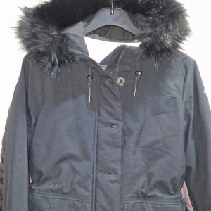 Svart parkajacka med päls från GANT - Parkajacka från GANT i svart med fluffig konstpäls runt huvan och quiltat vitt foder. Jackan har dragsko i midjan, stora fickor och både dragkedja och knappar framtill. Perfekt för kalla dagar med sin långa passform och varma material.