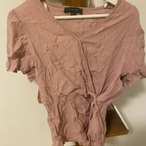 Rosa omlottklänning från Primark - Supersöt kort rosa klänning från Primark med omlottmodell och knyt i midjan. Klänningen har korta puffärmar och är tillverkad i ett lätt, skrynkligt bomullstyg som ger en avslappnad vibe. Perfekt för varma dagar när du vill känna dig både bekväm och snygg.