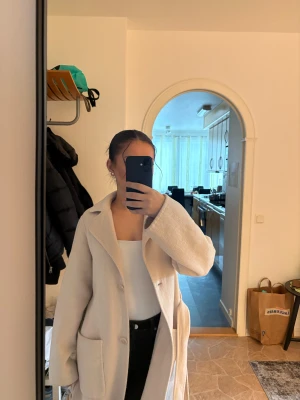 Beige kappa med stora fickor - Säljer en stilren beige kappa med stora fickor och klassisk krage. Kappan har knäppning framtill och en rak, loose passform som ger en chill vibe. Perfekt för dig som gillar minimalistisk stil och vill ha något som funkar till många outfits.