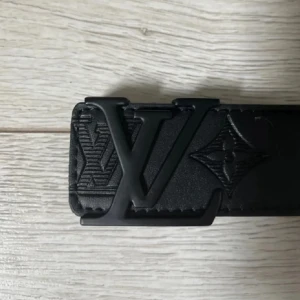 Svart Lou** Vui**** skärp i skinn - Säljer ett svart Lou** Vui**** bälte i skinn med präglade -loggor och monogramdetaljer längs hela remmen. Snygg, bred design med en stor -spänne i svart metall. Perfekt för dig som vill ha en lyxig och stilren accessoar med ikonisk känsla.