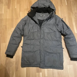 Grå pufferjacka med huva - Säljer en grå pufferjacka med huva och praktiska fickor framtill. Jackan har quiltad design, lång ärm med svarta muddar och dragkedja under frontslån. Perfekt för kalla dagar och har en clean, modern look.