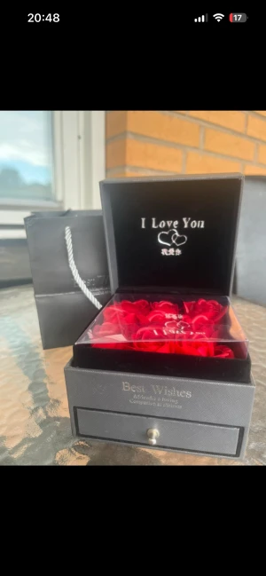 Svart smyckesask med röda rosor - Snygg svart smyckesask med texten 'I Love You' och två hjärtan på insidan locket. Inuti ligger röda rosor i plast, perfekt för att ge bort ett smycke som present. Kommer med matchande svart presentpåse med vit snodd. Boxen har en liten låda med metallknopp. Perfekt till en proposal 