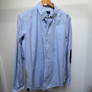 Ljusblå skjorta med svarta armbågslappar från Massimo Dutti - Snygg ljusblå skjorta från Massimo Dutti med klassisk button-down krage och svarta armbågslappar som ger en cool kontrast. Skjortan har långa ärmar och vita knappar framtill. Perfekt för dig som gillar stilrena detaljer och en modern look.