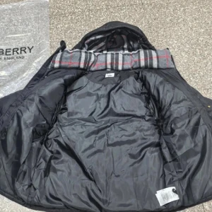 Svart pufferjacka från  - Svart pufferjacka från  med huva och klassiskt rutigt foder i grått, vitt och rött. Jackan har Burberry-logga på ärmen, dragkedja och knappar framtill. Perfekt för kalla dagar med sin tjocka och quiltade design.