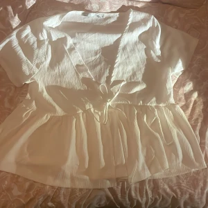Vit omlottblus från 157, storlek XL - Supersöt vit omlottblus från 157 i storlek XL. Blusen har korta ärmar, knytband i midjan och volangkant nedtill. Materialet är lätt och har en krinklad struktur som ger en chill och luftig känsla. Perfekt för vår och sommar!