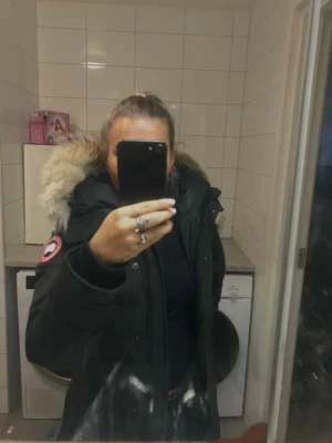 Svart lång dunjacka Canada Goose - Säljer en svart lång dunjacka från Canada Goose med ikonisk patch på ärmen och fluffig pälskantad huva. Jackan har dragkedja, knappar och två fickor framtill. Perfekt för kalla dagar och riktigt snygg passform.