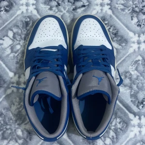 Nike Air Jordan 1 Low blå/vit/grå - Säljer ett par Nike Air Jordan 1 Low sneakers i blått, vitt och grått. Skorna har klassisk rund tå, snörning och grå swoosh på sidan. Ovandelen är i skinn och mesh, med perforerad tåbox för extra ventilation. Perfekta för dig som gillar streetwear och snygga detaljer.