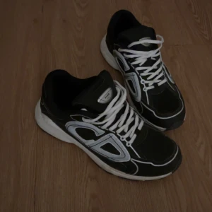 Vit/svarta skor  - Säljer ett par svarta sneakers med vita och grå detaljer samt tydlig CD-logga på sidan. Skorna har vita snören och en tjock, vit sula som ger en sportig vibe. Perfekta för dig som vill ha en clean och modern look.