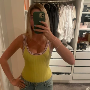 Gul ribbad body - Säljer en ljusgul ribbad body med breda axelband och djup rundad urringning. Toppen har en snygg randig kant i vitt, rosa och lila. 