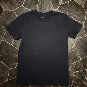  Hugo Boss Tshirt MörkBlå - Snygg svart t-shirt från Hugo Boss med diskret broderad logga på bröstet. Klassisk rund hals och korta ärmar. Tillverkad i mjuk bomull som känns skön mot huden. Perfekt för dig som gillar stilrena och enkla plagg med premiumkänsla.