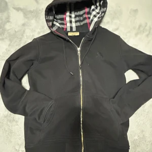 Svart Burberry hoodie med dragkedja - Svart hoodie från Burberry med klassiskt rutigt foder i huvan och snygg silverfärgad dragkedja framtill. Tröjan har snörning vid huvan, två fickor och långärmade ärmar. Perfekt för dig som gillar stilrena och exklusiva streetwear-plagg.