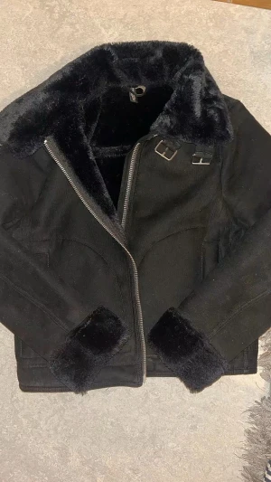 Svart pälsjacka med dragkedja  - Svart pälsjacka från H&M med fluffig fuskpäls på krage och ärmslut. Jackan har två snygga spännen vid halsen, silverfärgad dragkedja. Perfekt för dig som vill ha en cool och varm look i vinter.