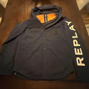 Mörkblå vindjacka från Replay - Snygg mörkblå vindjacka från Replay med stor vit logga längs ena ärmen och orange meshfoder på insidan. Jackan har huva, knappar framtill och två bröstfickor. Perfekt för dig som vill sticka ut med en sportig och modern look.