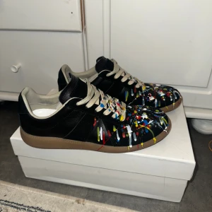Maison Margiela sneakers  - Unika svarta sneakers från Maison Margiela med färgstänk i rött, blått, gult och vitt över hela skon. Skorna har beige snörning, gummisula och är tillverkade i skinn. Perfekt för dig som vill sticka ut med en konstnärlig vibe.
