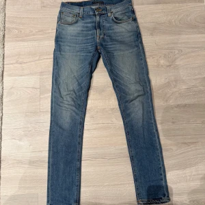 Nudie Jeans - Säljer ett par blå slim jeans från Nudie Jeans med klassisk femficksdesign och snygga slitningar. Jeansen har orange kontrastsömmar, läderpatch med logga bak och smal passform. Perfekt för dig som gillar stilrena och moderna jeans.
