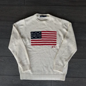 Vit stickad tröja Polo Ralph Lauren - Säljer en vit stickad tröja från Polo Ralph Lauren med amerikansk flagga på bröstet och RL-broderi. Klassisk rund halsringning och ribbade muddar. Perfekt för dig som vill ha en preppy och tidlös look.