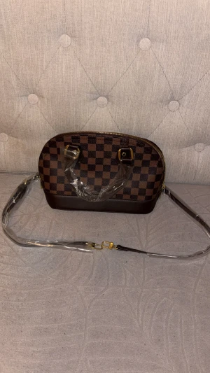 Axelväska Louis Vuitton - Snygg handväska från Louis Vuitton i klassiskt brunt rutigt mönster. Väskan har guldfärgade detaljer, avtagbar axelrem och dragkedja upptill. Insidan är beige med diskret LV-mönster och ficka. Perfekt storlek för det viktigaste och riktigt trendig look.