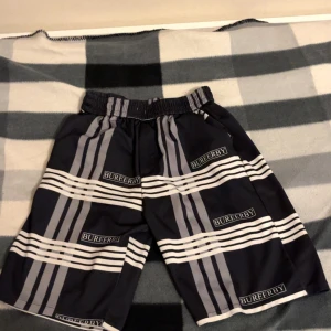 Randiga Bureerby shorts i svart och grå - Säljer ett par svarta och grå shorts från Bureerby med breda vita och grå ränder och logga tryckt över hela. De har elastisk midja och en loose passform, perfekta för chill dagar. Materialet känns syntetiskt och är lätt och bekvämt.