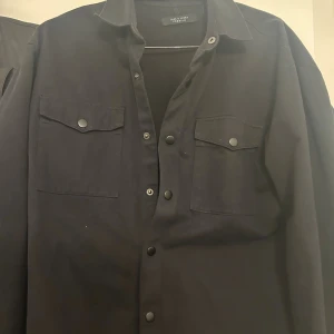 Svart overshirt från Jack & Jones - Svart overshirt från Jack & Jones med klassisk krage och tryckknappar framtill. Två stora bröstfickor med lock och knappdetaljer ger en cool look. Stl M
