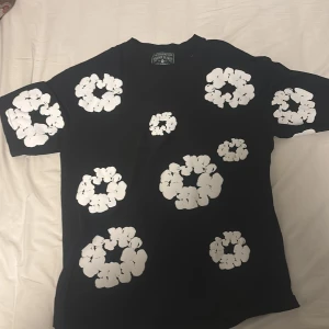 Svart t-shirt med vita blommor Denim Tears - Svart t-shirt från Denim Tears med stora vita blommönster tryckta över hela plagget, både fram och bak. Klassisk rund hals och korta ärmar. Materialet är mjuk bomull som känns skönt mot huden. Perfekt statement-plagg för dig som gillar unik design.