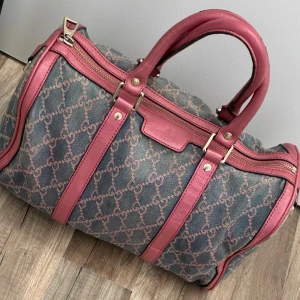 Gucci handväska med rosa detaljer - Gucci handväska i blått tyg med klassiskt GG-mönster och snygga rosa skinn-detaljer. Väskan har två handtag, guldfärgade dragkedjor och rymligt innerfack i beige bomull. Perfekt för dig som vill sticka ut med en ikonisk design.