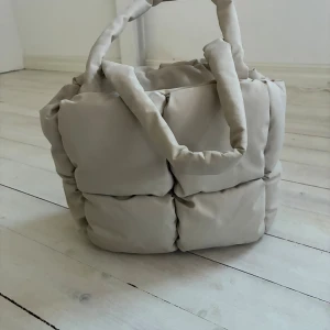 Beige quiltad axelväska i syntet - Supermjuk och fluffig beige axelväska med quiltad design. Väskan har breda, vadderade handtag och är tillverkad i syntetmaterial. Perfekt för dig som gillar trendiga accessoarer med unik form och cozy känsla.