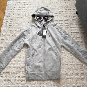 Grå hoodie med goggles från C.P. Company - Säljer en grå hoodie från C.P. Company med ikoniska goggles i huvan. Tröjan har hel dragkedja, två snedställda fickor med dragkedja och justerbar huva med snörning. Tillverkad i mjuk bomullsblandning och har ribbade muddar vid ärmslut och nederkant. Ny pris ligger på 2300 kr, därmed erbjuder jag ett generöst pris då den är helt ny oanvänt med lappar på 🙌😊