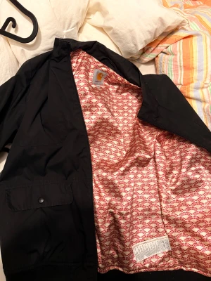 Svart  Carhartt Jacka - Svart bomberjacka från Carhartt med coolt mönstrat innerfoder i rött och vitt. Jackan har dragkedja framtill, två fickor med lock och knappar, samt ribbade muddar vid midja och ärmslut. Perfekt för dig som gillar streetwear och snygga detaljer.