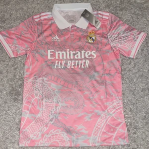 Real Madrid rosa drakmönstrad tröja - Säljer en sjukt snygg Real Madrid fotbollströja som är rosa med grått drakmönster. Tröjan har vit krage, korta ärmar och klassiska Adidas-ränder på axlarna. Materialet är lätt och andas, perfekt för träning eller match. Emirates Fly Better tryck på bröstet.